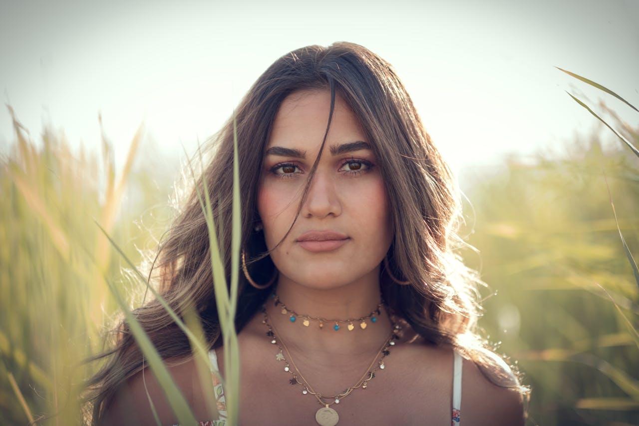 A beautiful young woman posing in a sunny field, wearing necklaces and a floral dress.