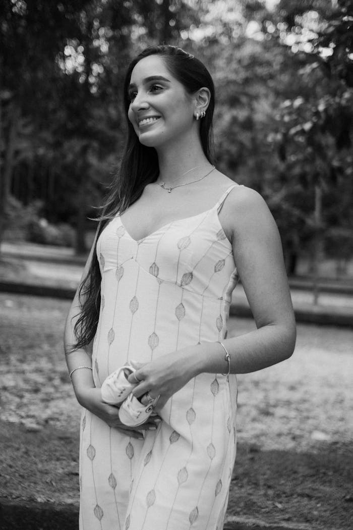 Smiling pregnant woman holds baby shoes, standing outdoors during the day.